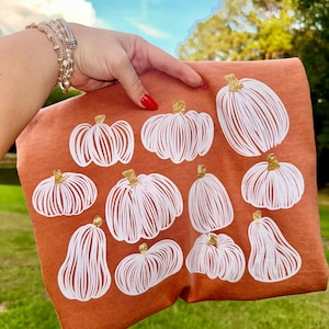 Può includere: T-shirt color ruggine con un motivo di zucche bianche con steli dorati. Le zucche variano per forma e dimensione, creando un design a tema autunnale. La maglietta è tenuta contro uno sfondo di erba verde e cielo blu.