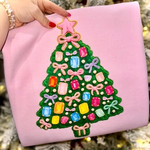 May include: A light pink sweatshirt featuring a Christmas tree design. The green tree is adorned with colorful gift-shaped ornaments, pink bows, and a pink star at the top. The design is embroidered with gold thread and pearls.