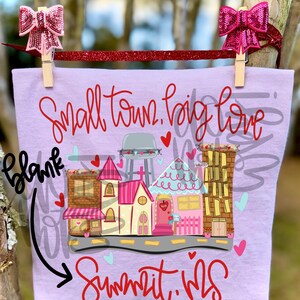 May include: A light purple t-shirt with the text "Small town, big love" and "Summit, MS" in red script. The shirt features a colorful graphic of a town with buildings, a water tower, and hearts. Two pink sequined bows are clipped to the top.