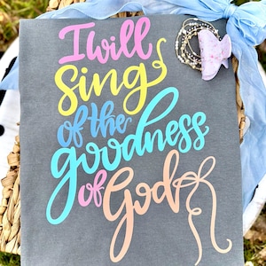 May include: A gray fabric sign with the text "I will sing of the goodness of God" in colorful script. A light blue ribbon and a bracelet with a butterfly accent are attached.