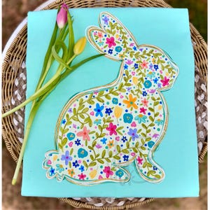 May include: A light blue square fabric with a floral bunny design. The bunny is filled with colorful flowers and green leaves, outlined in gold. A few tulips in pink and yellow are placed on the left side of the fabric.