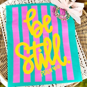 以下が含まれることがあります： 垂直のピンクのストライプが入った、ターコイズ色のTシャツ。「be still」の文字は黄色の筆記体で、下に「PSALM 46:10」と書かれています。ピンクのリボン、ブレスレット、ヘアクリップが右上にあります。