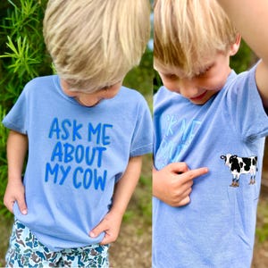 Könnte beinhalten: Zwei hellblaue T-Shirts mit dem Text "ASK ME ABOUT MY COW" und einer Kuhgrafik. Die Shirts werden von einem Kind getragen. Die Kuhgrafik ist schwarz-weiß mit braunen Stiefeln.