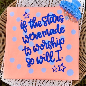 Op de afbeelding: Koraalrood T-shirt met de blauwe tekst "if the stars were made to worship so will I". Het shirt heeft lichtblauwe stippen en steraccenten. Een lichtblauwe bloemen haarclip en kralenarmbanden bevinden zich rechtsboven.