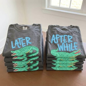 May include: Piles of grey t-shirts with the words "LATER" and "AFTER WHILE" in light blue. Each shirt has a green alligator design with red details. The shirts are folded and stacked on a brown surface.