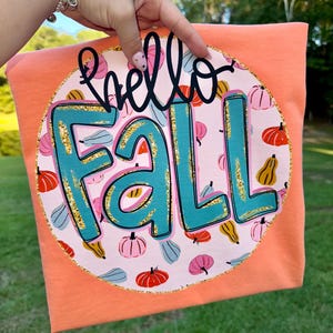 Puede incluir: Camiseta color melocotón con un diseño circular con las palabras "hello FALL" en negro y turquesa. El diseño incluye calabazas de varios colores y un borde dorado con purpurina.