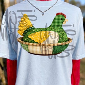 May include: Light blue t-shirt featuring a whimsical hen design. The hen is crafted from green gingham and yellow polka dot fabric, nestled in a basket. The design includes a red comb and a yellow beak. The shirt is worn over a red top.