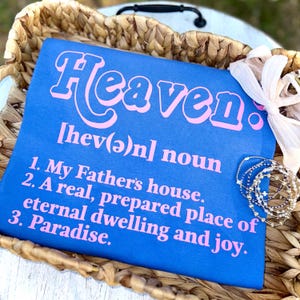 May include: A blue t-shirt with the word "Heaven" in pink, along with a definition. The shirt is in a woven basket with bracelets and a decorative flower. The definition includes "My Father's house" and "Paradise."