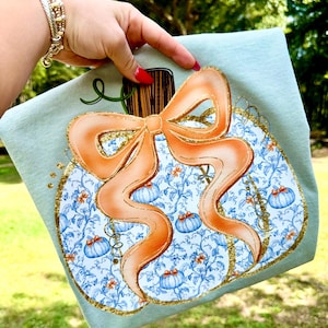 Könnte beinhalten: Ein hellblaues Dekoreck mit einem Kürbismotiv. Der Kürbis hat eine große orangefarbene Schleife, goldene Glitzerakzente und ein blau-weißes Blumenmuster. Kleine blaue Kürbisse sind über das Design verstreut.