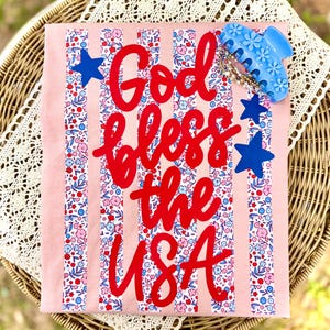 May include: A light pink t-shirt with the text "God Bless the USA" in red script. The shirt features vertical stripes with a floral pattern in red, white, and blue. Blue star accents and a blue hair clip are on top.