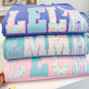 May include: Three stacked, folded t-shirts in shades of blue, teal, and pink. Each shirt features a name spelled out in large, floral-patterned letters with scalloped edges. The letters have a mix of polka dots and floral designs.