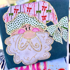 May include: A teal sweatshirt featuring a Santa Claus design. The Santa has a pink and white hat with red bows and green accents. His face is pink with a white beard outlined in gold glitter. A large green bow with white polka dots is on the side.