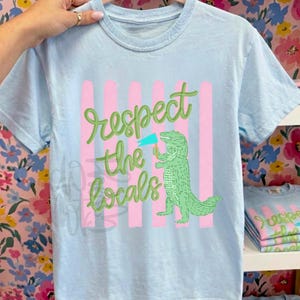 May include: Pale blue t-shirt featuring the phrase "Respect the Locals" in green script. The design includes a green alligator holding a flag, set against pink vertical stripes. The t-shirt is displayed on a rack.