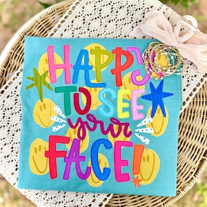 Könnte beinhalten: Ein türkisfarbenes T-Shirt mit dem Schriftzug "HAPPY TO SEE your FACE!" in bunten, geschichteten Buchstaben. Das Shirt hat gelbe Smiley-Gesichter und Sternakzente. Eine Sammlung von Armbändern und eine dekorative Schleife liegen obenauf.