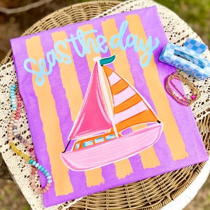 Op de afbeelding: Een paars t-shirt met oranje verticale strepen en een zeilbootafbeelding. Het shirt heeft de tekst "Seas the day" in lichtblauw. Een blauw-wit geruite haarclip en kralenkettingen zijn ook zichtbaar.