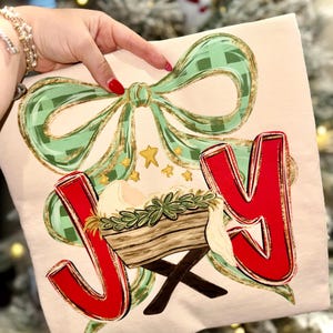 May include: A cream-colored t-shirt with a Christmas-themed design. The design includes a green plaid bow with gold accents, the word "JOY" in red, and a depiction of the nativity scene. Gold stars are also included in the design.