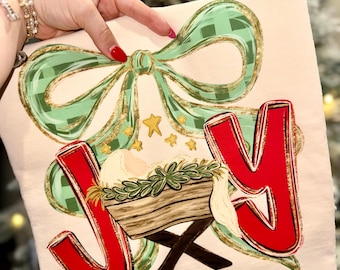 May include: A cream-colored t-shirt with a Christmas-themed design. The design includes a green plaid bow with gold accents, the word "JOY" in red, and a depiction of the nativity scene. Gold stars are also included in the design.