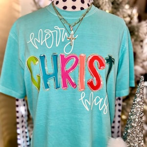 May include: A turquoise t-shirt with the words "Merry Christmas" in white script and the word "CHRISTMAS" in colorful block letters. The shirt is adorned with a gold cross pendant necklace.