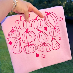 Puede incluir: Sudadera rosa claro con un patrón repetido de contornos de calabazas rosas y diamantes. Las calabazas varían en tamaño y estilo, creando un diseño otoñal. La sudadera es sostenida por una mano.