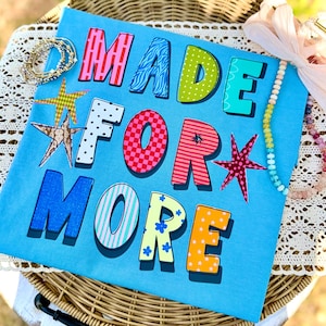 Könnte beinhalten: Ein hellblaues T-Shirt mit den Worten "MADE FOR MORE" in bunten, gemusterten Buchstaben. Dekorative Sterne und Schmuck sind um das T-Shirt herum angeordnet, das auf einem Weidenkorb präsentiert wird.