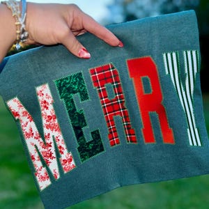 Pu&ograve; includere: Un oggetto in tessuto color verde acqua con la scritta "MERRY" applicata in vari motivi. Le lettere sono in rosso, verde e bianco con disegni floreali, a quadri e a righe. L'oggetto &egrave; tenuto da una mano.
