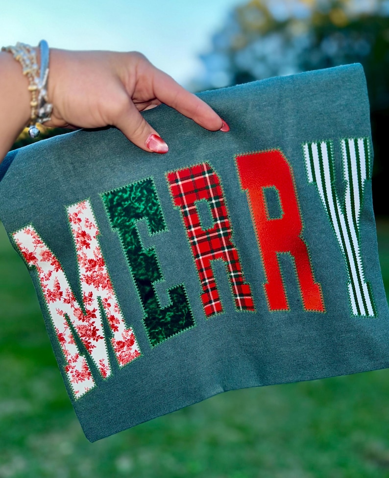 Pu&ograve; includere: Un oggetto in tessuto color verde acqua con la scritta "MERRY" applicata in vari motivi. Le lettere sono in rosso, verde e bianco con disegni floreali, a quadri e a righe. L'oggetto &egrave; tenuto da una mano.