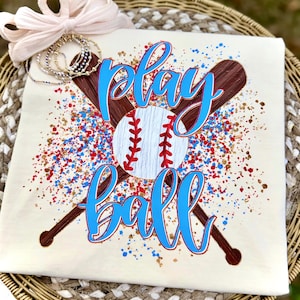 May include: Cream-colored t-shirt with a baseball theme. The design features a baseball, crossed bats, and the words "play ball" in blue script. Red, blue, and gold paint splatters add a decorative touch. A bow and beaded bracelets are in the background.