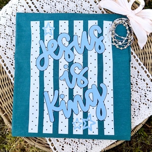 以下が含まれることがあります： 水色のTシャツ。明るい青色で「Jesus is King」というフレーズが書かれており、濃い青色で縁取られています。デザインには白い縦縞、黒い水玉模様、明るい青色の星のアクセントが含まれています。Tシャツは籐の表面に表示されています。