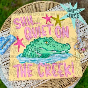 May include: A light yellow t-shirt featuring a green alligator graphic in water with pink stars and the text SHH... QUIET ON THE CREEK! in pink lettering. The design includes a yellow star and a light blue star accent. The shirt is displayed on a wicker surface.