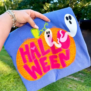 May include: A blue Halloween-themed t-shirt featuring a large orange pumpkin with a leopard print design. The word "HALLOWEEN" is written in bright pink letters. Two cartoon ghosts and a bat are also featured.