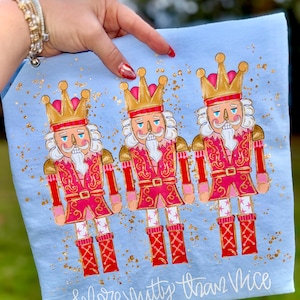 May include: A light blue decorative towel featuring three cartoon nutcracker figures in red and pink uniforms with gold crowns. The towel has gold glitter accents and the text "More Nutty Than Nice" in white script.