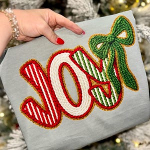 May include: A gray fabric item with the word "JOY" embroidered in red, white, and green yarn. The letters are outlined in gold, and a green yarn bow is at the top right. The item is held by a person with red nail polish.