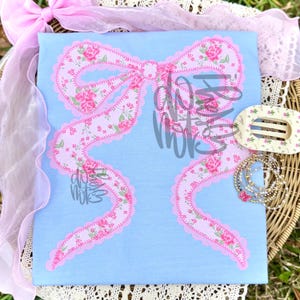 May include: A light blue t-shirt featuring a large pink bow design with a floral pattern and scalloped edges. The bow has long, flowing ribbons. A matching hair clip and bracelets are also visible.