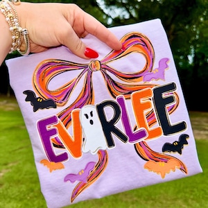 May include: A lilac t-shirt with the name "EVRLEE" in orange, black, and purple, with ghost and bat graphics. The shirt features a large bow design with orange and purple accents, ideal for Halloween.