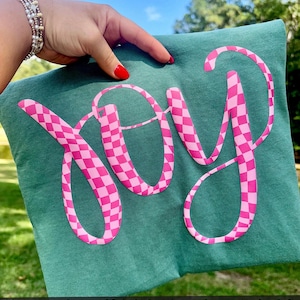 Puede incluir: Un cojín verde azulado con la palabra "JOY" en letras grandes, rosas y a cuadros. El cojín está sostenido por una mano con esmalte de uñas rojo. El texto en la parte inferior dice "Please note this is puff vinyl."