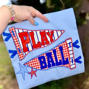 May include: Light blue t-shirt with two pennant designs. The top pennant reads "PLAY" in red block letters, and the bottom pennant reads "BALL" in blue. Both pennants have a red and white checkered pattern and are decorated with blue ribbons and stars.