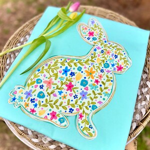 May include: A light blue decorative piece featuring a floral bunny design. The bunny is filled with colorful flowers and green vines, outlined in gold. A sprig of tulips rests on the side, adding a touch of spring.
