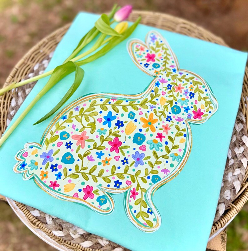 May include: A light blue decorative piece featuring a floral bunny design. The bunny is filled with colorful flowers and green vines, outlined in gold. A sprig of tulips rests on the side, adding a touch of spring.