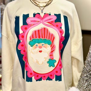 May include: Cream-colored sweatshirt featuring a Santa Claus graphic. The Santa design is framed in pink with a bow at the top and a scalloped edge. Santa wears a red and pink striped hat and has a white beard. The background has navy blue vertical stripes.