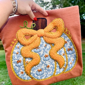 May include: An orange-colored decorative pillow featuring an embroidered pumpkin design. The pumpkin has a blue and white floral pattern with orange accents and a large, textured orange bow. The pillow is held by a person.