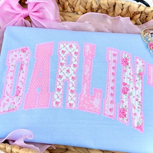 May include: A light blue t-shirt with the word "DARLING" in pink, floral-patterned letters. The letters are outlined in a darker pink and have a stitched appearance. A pink bow and jewelry are in the background.