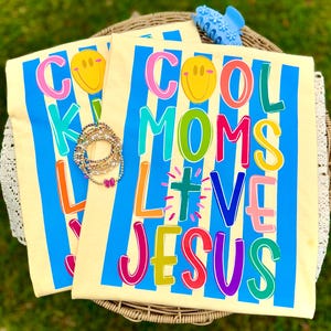 Puede incluir: Dos camisetas amarillo claro con el texto "COOL MOMS LOVE JESUS" en letras coloridas y una carita sonriente. Las camisetas tienen rayas verticales azules. También se ve una colección de pulseras y una pinza para el pelo azul.