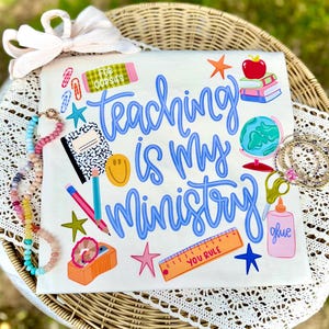 May include: White t-shirt with the phrase "Teaching is My Ministry" in blue script. The shirt is decorated with school-themed graphics, including an apple, books, a globe, and pencils. Additional graphics include stars, a smiley face, and a ruler.