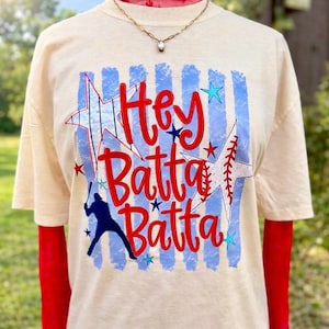 May include: Cream-colored t-shirt with the words "Hey Batta Batta" in red script. The design includes blue vertical stripes, stars, and a baseball bat and player silhouette. The shirt has short sleeves.