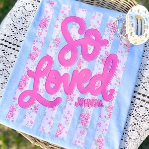 May include: A light blue decorative pillow with the words "So Loved" in bright pink script. The pillow has a floral pattern with pink roses and vertical stripes. The text "John 3:16" is below the word "Loved".
