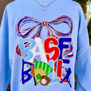 May include: Light blue sweatshirt with a baseball theme. The word "BASEBALL" is displayed in a colorful, layered design. A large bow graphic is at the top. Baseball bat, ball, glove, and player silhouette complete the design.