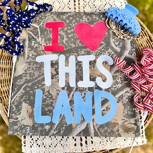 Può includere: Una tela quadrata con un motivo mimetico e le parole "I LOVE THIS LAND" in rosso, bianco e blu. Elementi decorativi, tra cui un fermaglio blu e nastro rosso e bianco, circondano la tela.