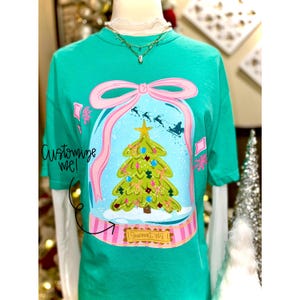 May include: A teal t-shirt with a snow globe design. It features a Christmas tree, snowflakes, and a pink bow. The text "Customize me!" is on the left. The bottom of the globe has the text "Sumner, WA".