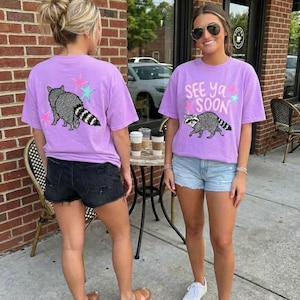 May include: Two women wearing lilac t-shirts featuring a raccoon design and the words "See Ya Soon". The t-shirts have a crew neck and short sleeves. The raccoon is grey and white with a striped tail. The shirts are worn with denim shorts.