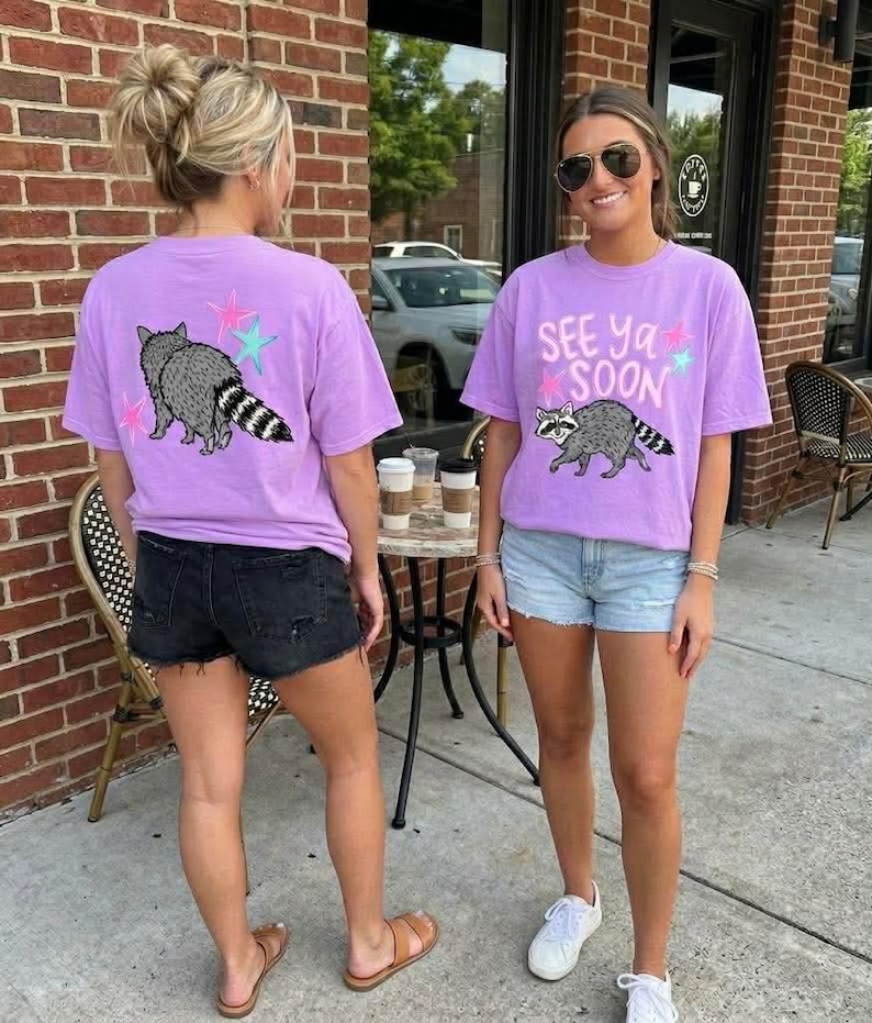 May include: Two women wearing lilac t-shirts featuring a raccoon design and the words "See Ya Soon". The t-shirts have a crew neck and short sleeves. The raccoon is grey and white with a striped tail. The shirts are worn with denim shorts.
